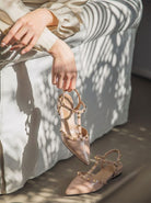 Moncelia beige studded T-strap flats, held by seated model showing the T-strap, women's sizes 5 to 10.5